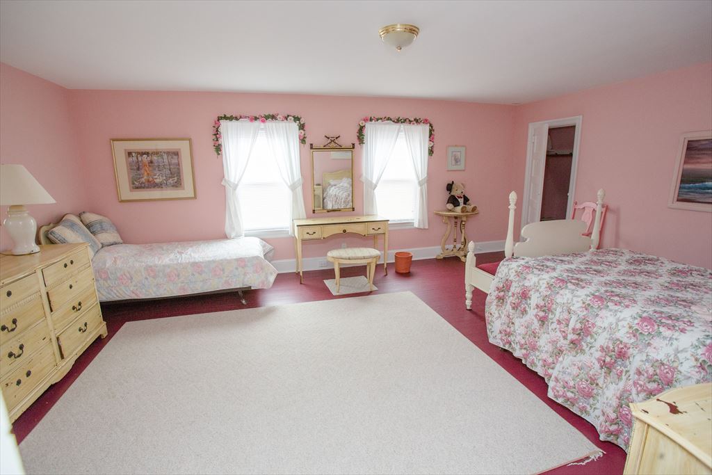 Large pink bedroom with two beds, light wood dressers, a vanity with mirror and rose-garland window valances